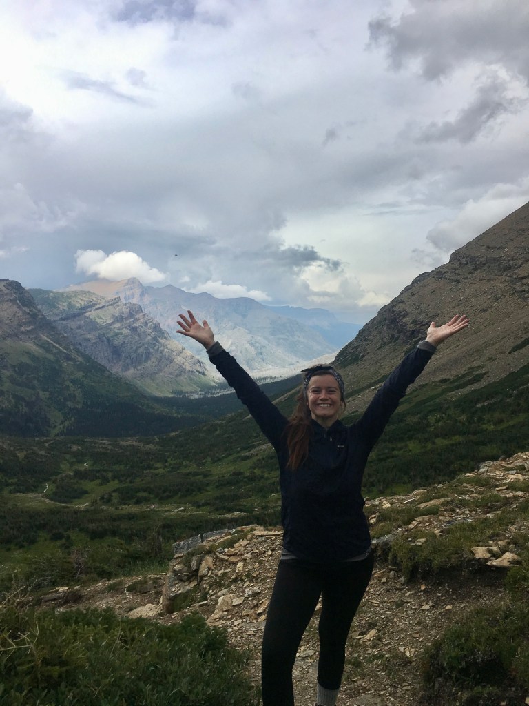 A picture of McKenzie smiling, with her arms upraised. Mountains are in the background.