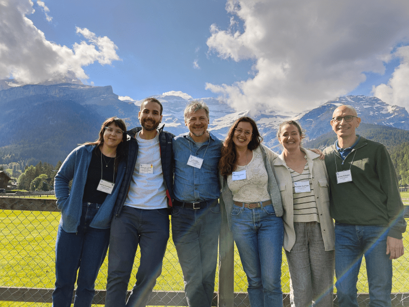 A group of 6 current and former lab members are smiling and standing close together. There are snow-capped mountains in the background.