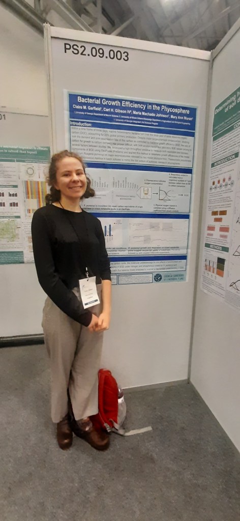 Claire Garfield stands smiling in front of a corner with posters on the wall.