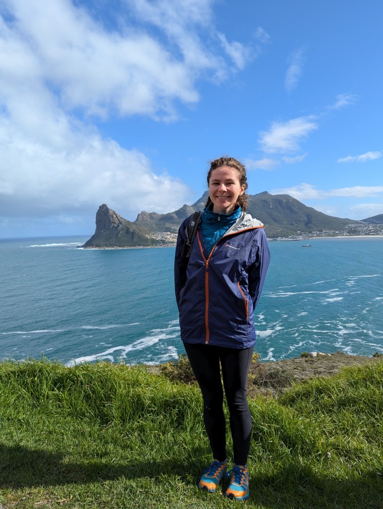Claire Garfield stands smiling in the grass. The ocean and an island are in the background. 